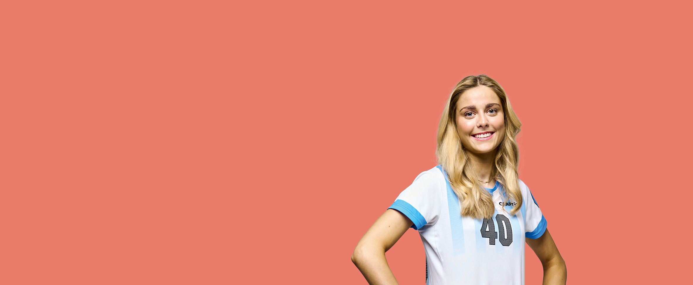 En ung, smilende kvinde med lyst hår står med hænderne i siden mod en ensfarvet koralrød baggrund. Hun er iført en hvid sportsbluse med lyseblå kanter og tallet 40 trykt på brystet.