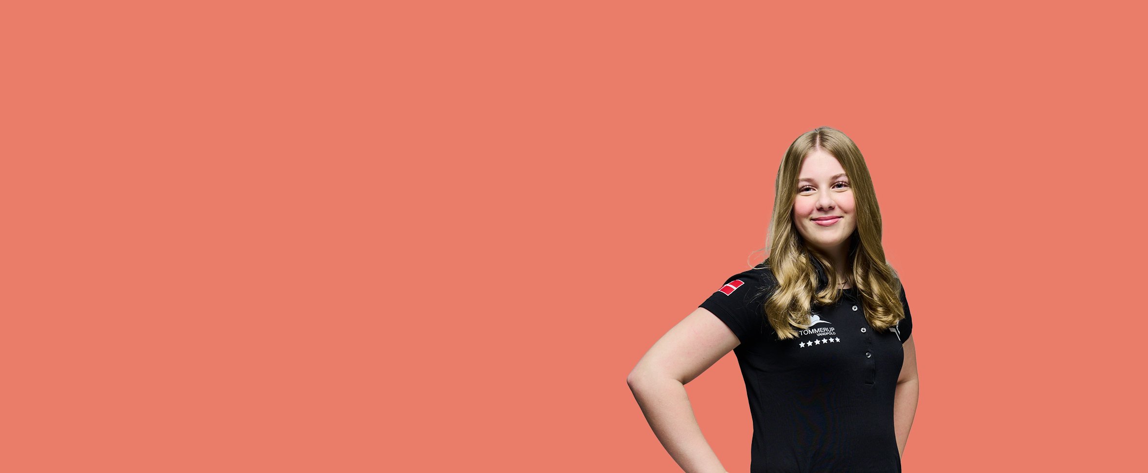 Portræt af en smilende ung kvinde med langt lyst hår, iført en sort t-shirt med logoer, der poserer med hænderne i siden mod en ensfarvet laksefarvet baggrund.