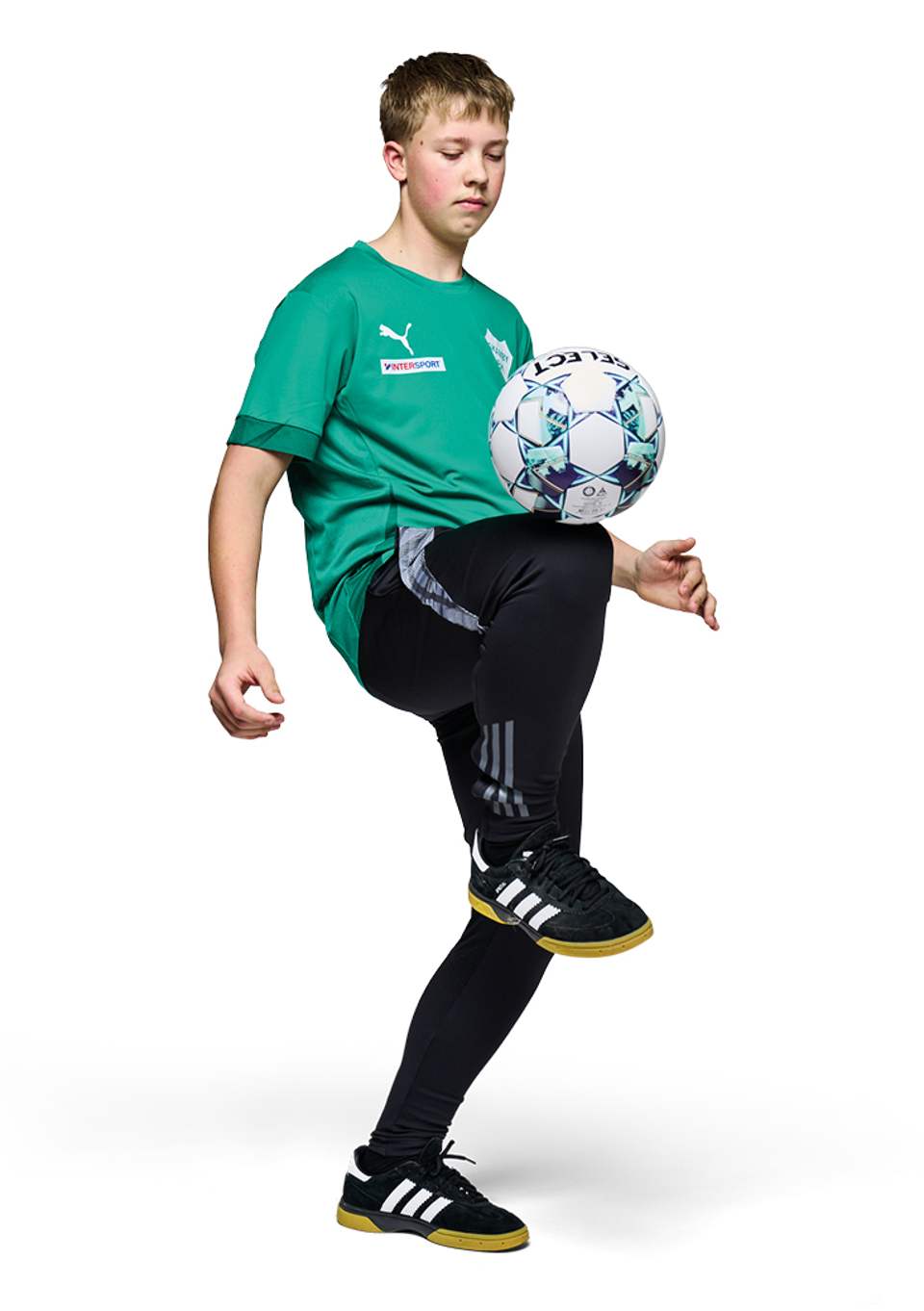 En ung dreng i en grøn sportstrøje og sorte træningsbukser jonglerer koncentreret med en fodbold på sit højre knæ, mens han balancerer på det venstre ben mod en hvid baggrund.