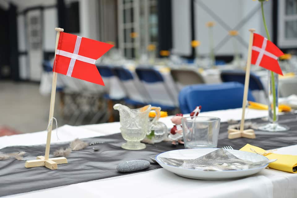 Et festligt dækket bord med hvid dug, en grå løber og små dannebrogsflag på træstativer. På bordet ses også service, glas og pynt som fjer og sten. I den slørede baggrund anes flere dækkede borde med blå stole.