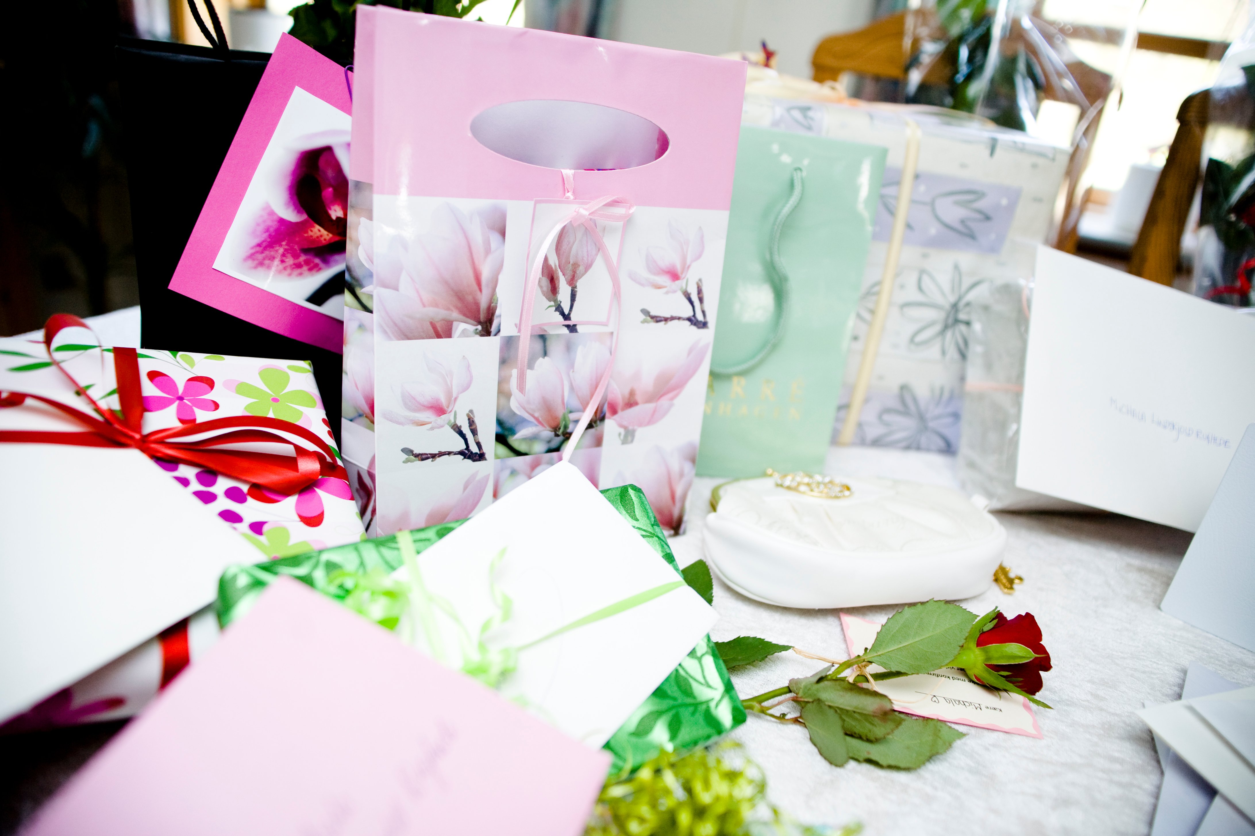 Et udvalg af gaver, gaveposer og lykønskningskort samlet på et bord. Der ses blandt andet en lyserød gavepose med blomstermotiver, en pakke indpakket i blomstret papir med et rødt bånd, en lille hvid taske og en enkelt rød rose.