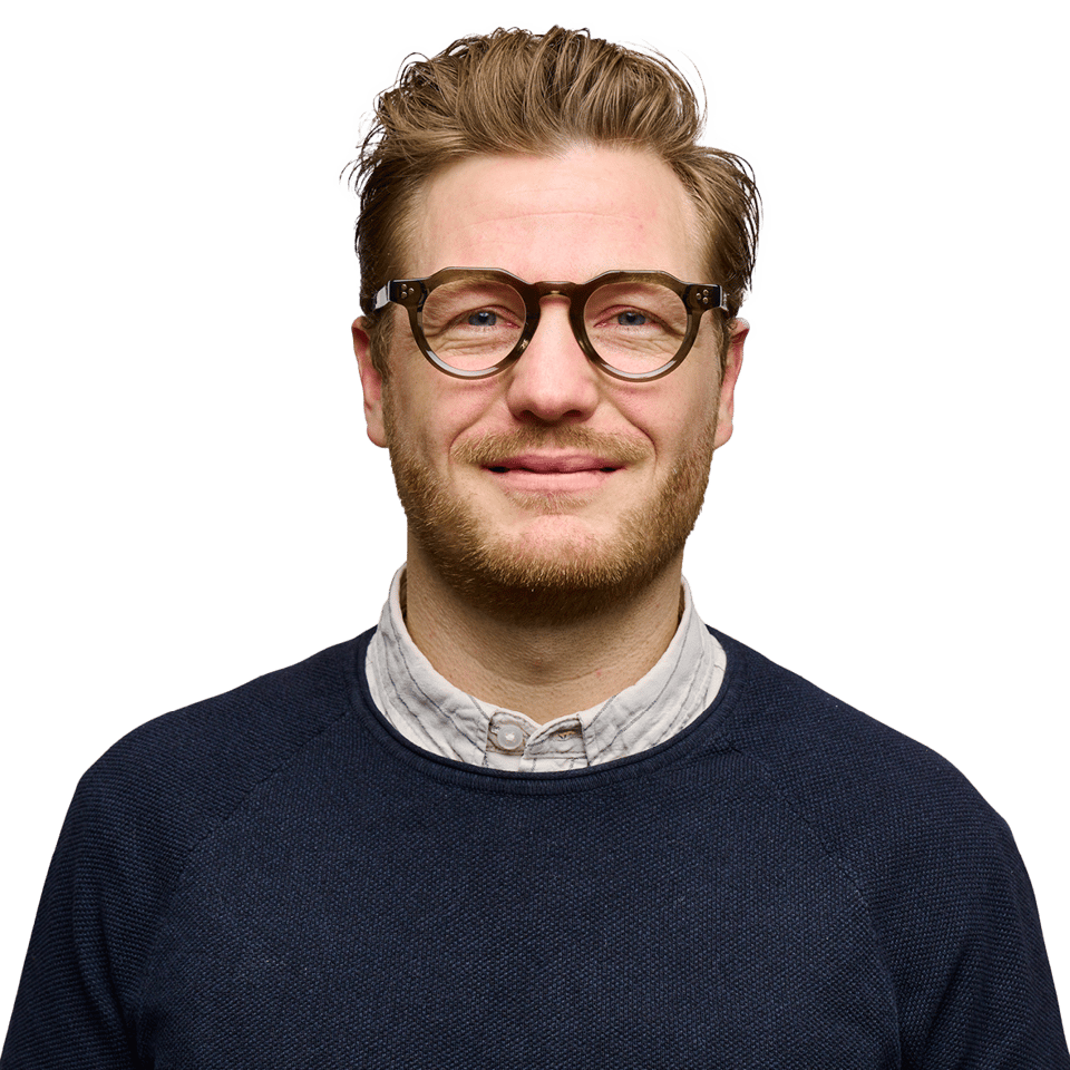 Portrætfoto af en smilende mand med lyst, tilbagestrøget hår, briller og kort skæg, iført en mørkeblå sweater over en lys skjorte.