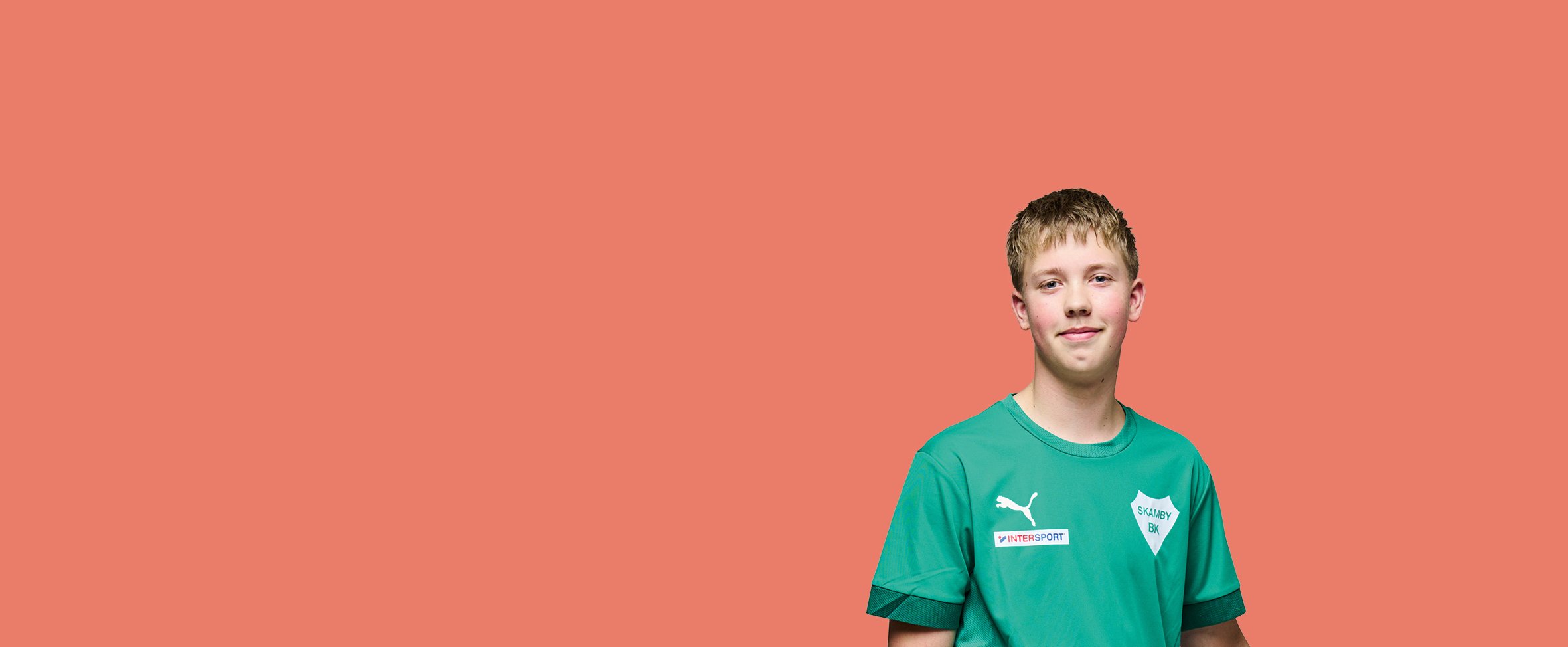 En ung mand med lyst hår smiler svagt mod kameraet. Han er iført en grøn Viborg FF-sportstrøje og er placeret til højre i billedet mod en ensfarvet koralfarvet baggrund.