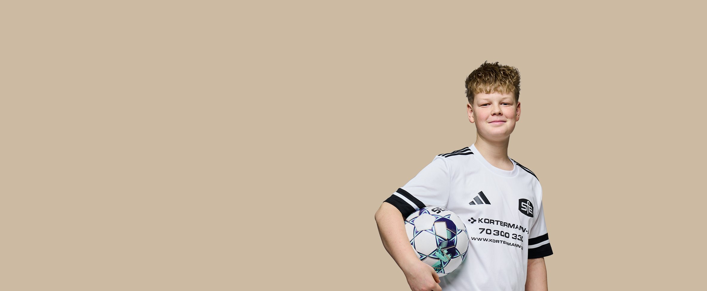 Portræt af en smilende ung dreng i en hvid fodboldtrøje, der holder en fodbold under armen mod en neutral baggrund.