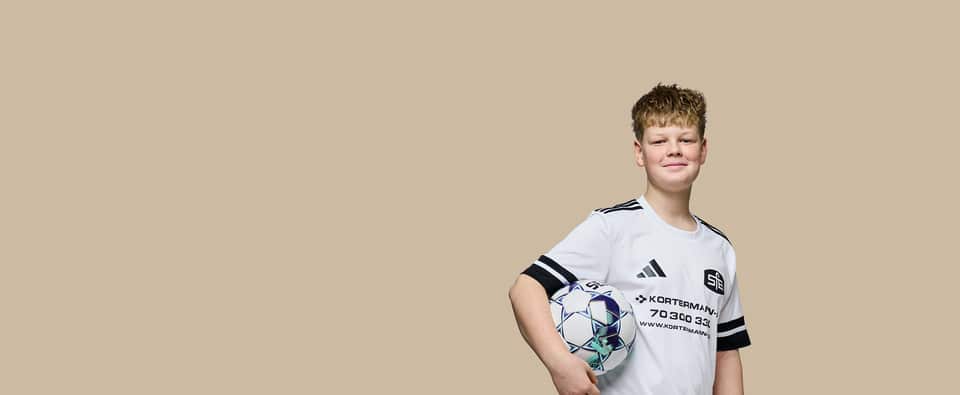 Portræt af en smilende ung dreng i en hvid fodboldtrøje, der holder en fodbold under armen mod en neutral baggrund.