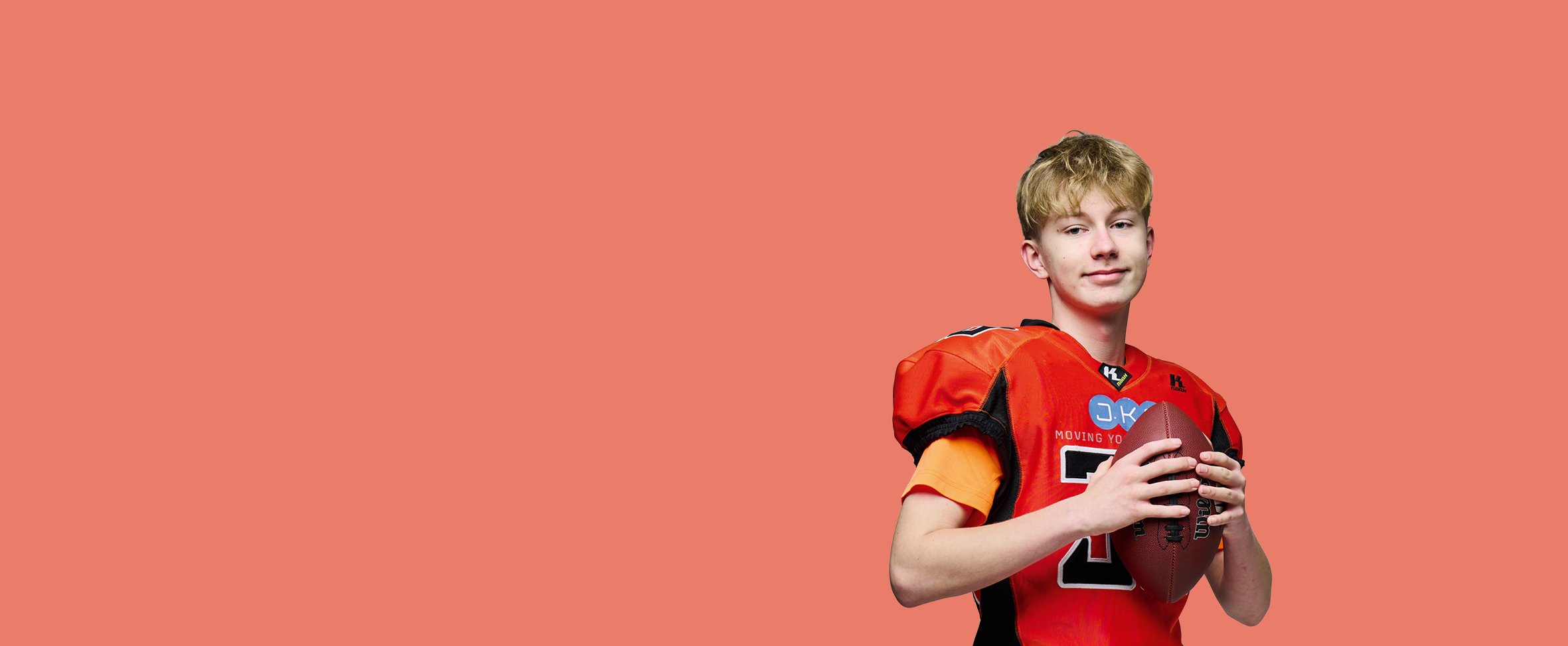 En ung, lyshåret mand i en rød og sort amerikansk fodboldtrøje med skulderpuder holder en amerikansk fodbold foran en ensfarvet koralfarvet baggrund.
