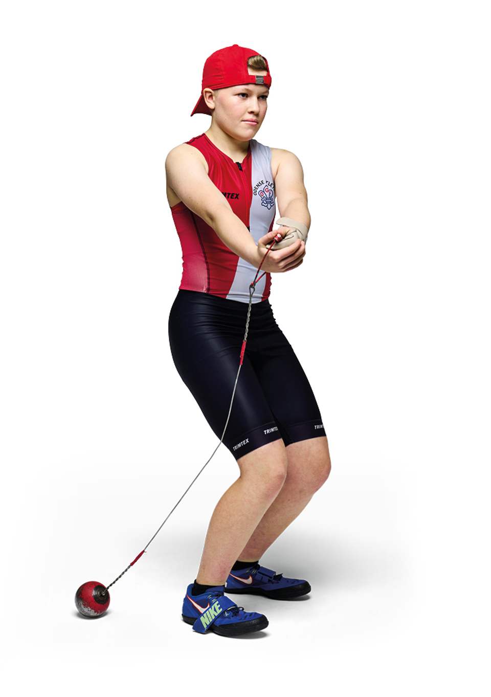 En ung atlet i fuld figur, der forbereder et hammerkast mod en hvid baggrund. Hun er iført en bagvendt rød kasket, en rød og grå top, sorte sportsshorts og blå atletiksko. Atleten står i en koncentreret position med armene strakt nedad og hænderne samlet om hammerens håndtag, mens metalkuglen hviler på jorden bag hende.