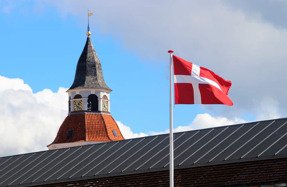 Et dansk flag, Dannebrog, der vajer i vinden ved siden af et kirketårn med et mørkt spir og en urskive. I forgrunden ses et mørkt, rillet tag, og i baggrunden er der en blå himmel med hvide skyer.