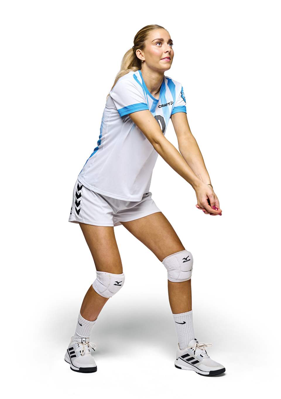 En blond kvindelig volleyballspiller i en hvid og lyseblå spillertrøje og hvide shorts står i en modtagelsesposition med samlede, strakte arme. Hun har hvide knæbeskyttere og sportssko på og kigger opad mod en hvid baggrund.