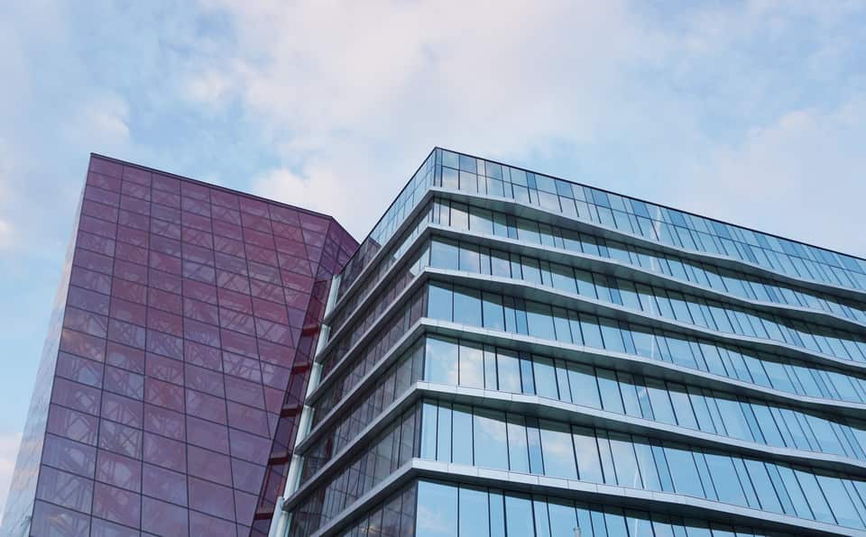 Lavvinkelbillede af en moderne glasbygning mod en lys himmel. Til venstre ses en sektion med mørkelilla glas i et gittermønster, og til højre en større facade med vandrette vinduespartier og hvide, zigzag-formede bånd.