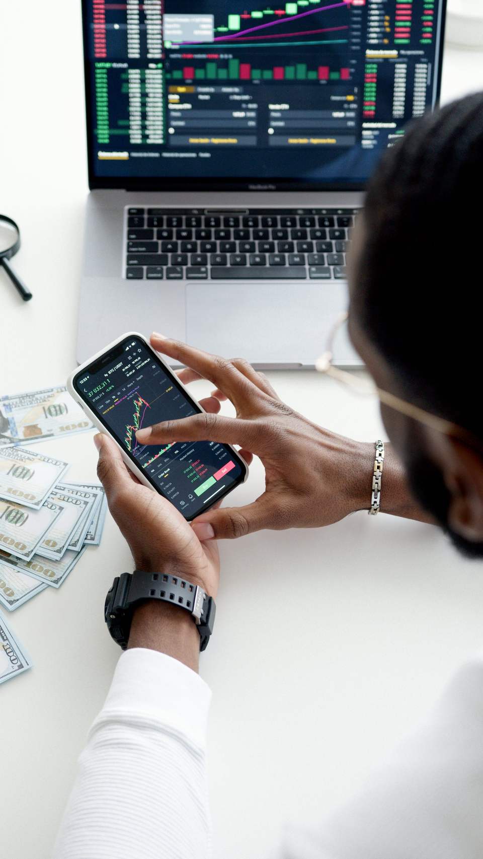 En person ses bagfra over skulderen, mens vedkommende analyserer finansielle grafer og børskurser på en smartphone. På et hvidt skrivebord står en bærbar computer, der også viser handelsdata, og flere 100-dollarsedler ligger spredt ved siden af telefonen.