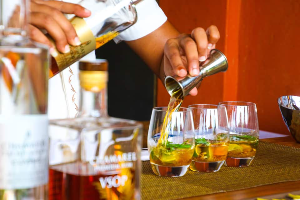 Nærbillede af en bartender, der hælder en gylden væske fra et målebæger i et af tre glas med cocktails indeholdende mynteblade og is. I forgrunden ses flasker med alkohol, herunder en mærket VSOP.