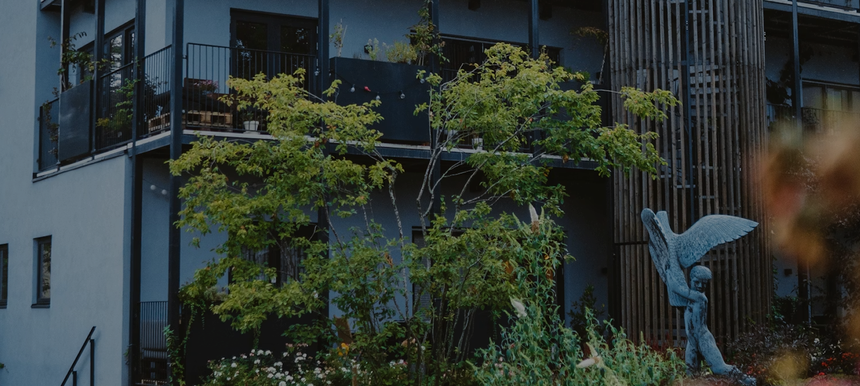 Moderne facade af en bygning med altaner prydet med planter, et lysegrønt træ i forgrunden og en skulptur af en bevinget skikkelse ved siden af en rund bygningsdel med lodrette trælameller.