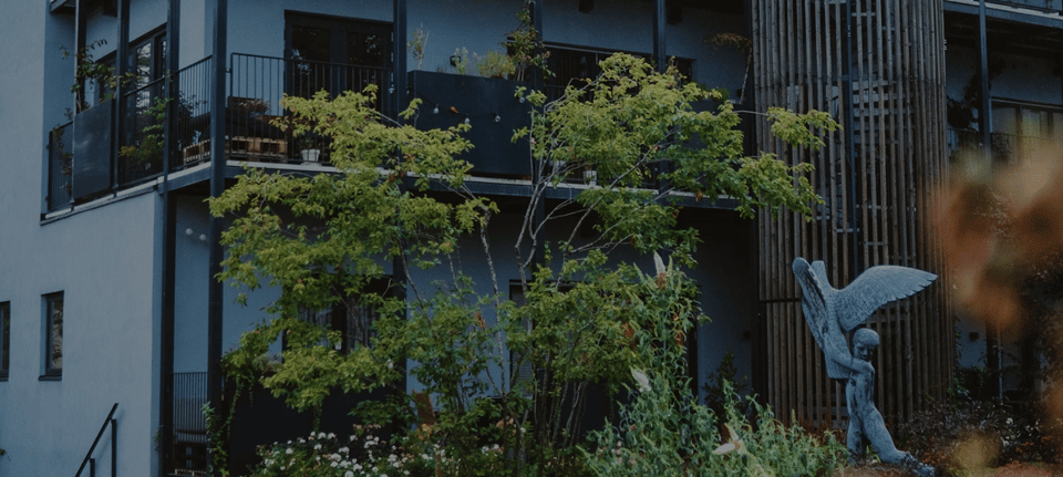 Moderne facade af en bygning med altaner prydet med planter, et lysegrønt træ i forgrunden og en skulptur af en bevinget skikkelse ved siden af en rund bygningsdel med lodrette trælameller.