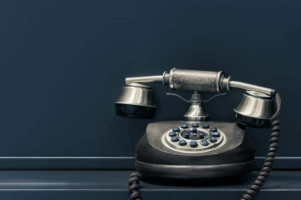 Antik-inspireret sort og sølvfarvet telefon med trykknapper arrangeret som en drejeskive, placeret på en mørkeblå overflade mod en mørk baggrund.