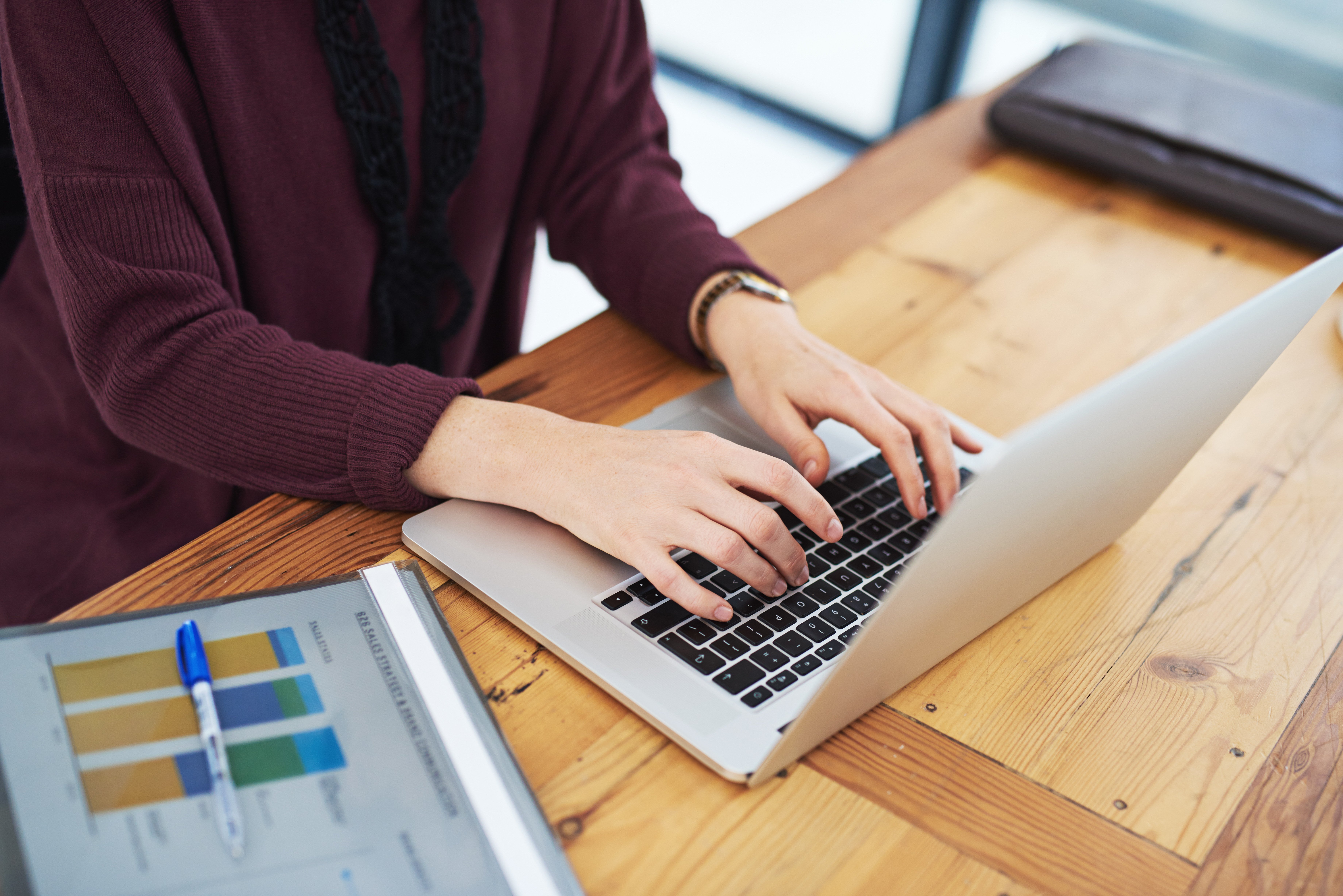 En person i en lilla trøje skriver på en bærbar computer, der står på et lyst træbord. I forgrunden ligger et dokument med et farverigt søjlediagram og en blå kuglepen.