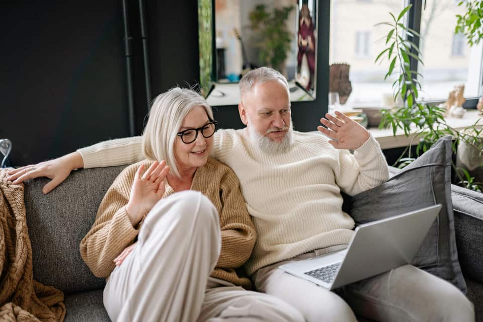 Et ældre par sidder sammen i en sofa og vinker smilende til en bærbar computer under et videoopkald i en hyggelig stue med planter i baggrunden.